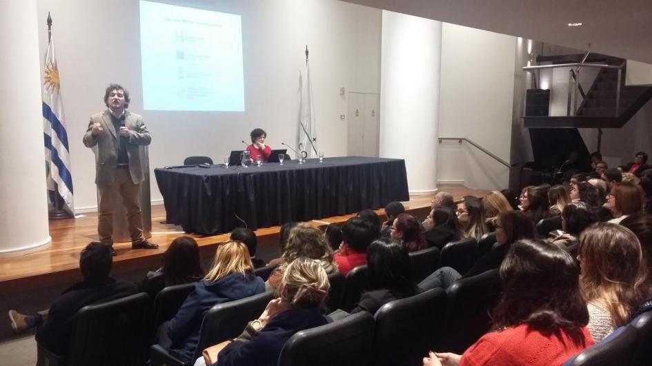 Salón de eventos con gran marco de público, persona presentando en estrado. 