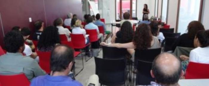 Mujer parada de frente hablando a un grupo de personas sentadas en una sala