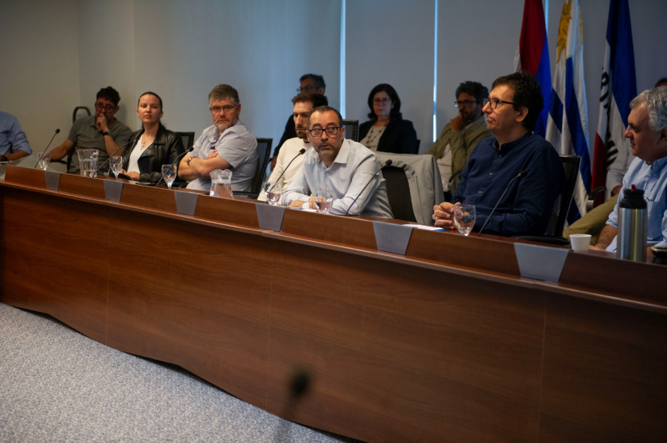 Panel de siete personas en discusión profesional con micrófonos y agua sobre la mesa.