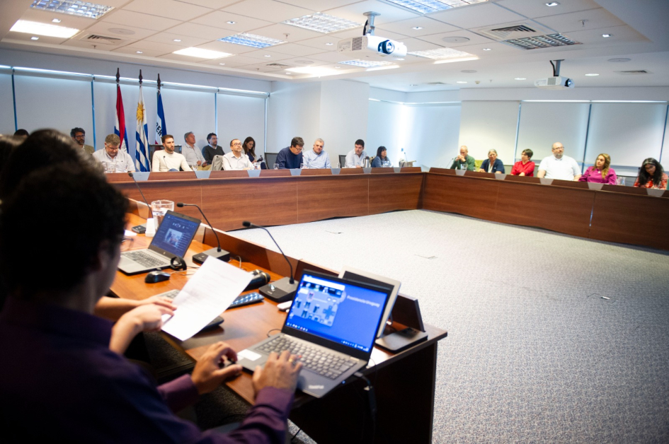 Mesa en U con asistentes en discusión profesional; banderas y proyectores en sala iluminada.