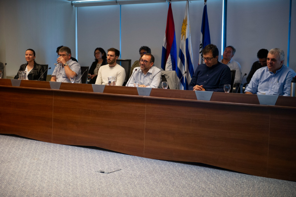 Panel de siete personas en discusión profesional con micrófonos y agua sobre la mesa.