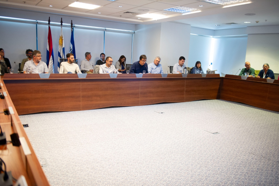 Mesa en U con asistentes en discusión profesional; banderas y proyectores en sala iluminada.