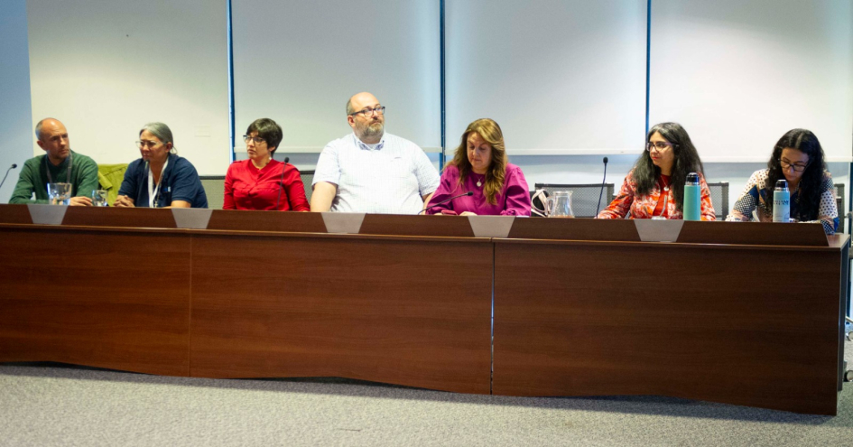 Panel de siete personas en discusión profesional con micrófonos y agua sobre la mesa.