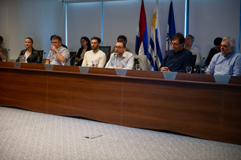 Panel de siete personas en discusión profesional con micrófonos y agua sobre la mesa.