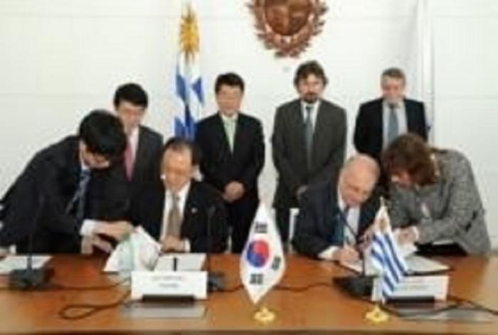 Corea Uruguay firma 2013 en mesa con banderas de cada país dos hombres firmando, rodeados de autoridades y asistentes.