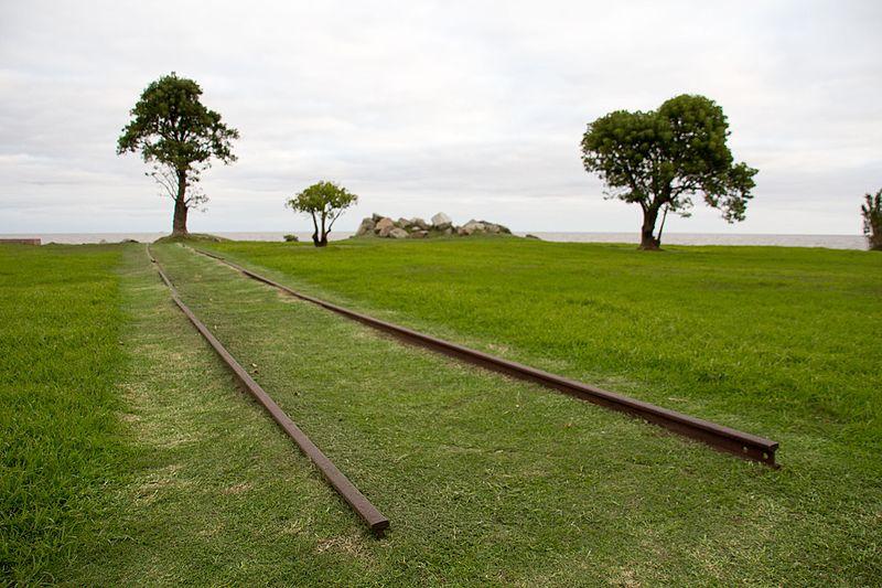 Memorial al Holocausto del Pueblo Judio – Fotografía de Mike Peel – www.mikepeel.net – CC-BY-SA-4.0 Fotografía que muestra un área verde, con referencia a unos trozos de vías de tren. Contra el horizonte, la silueta de tres árboles.