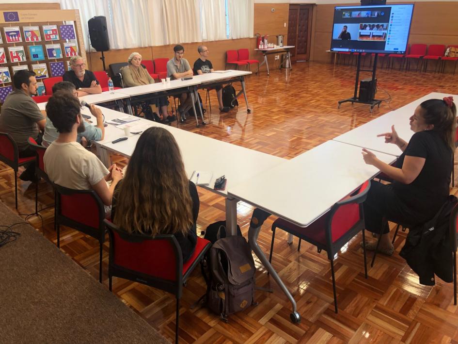 Grupo de trabajo reunido alrededor de una mesa, frente a este, televisor con conectados por zoom.