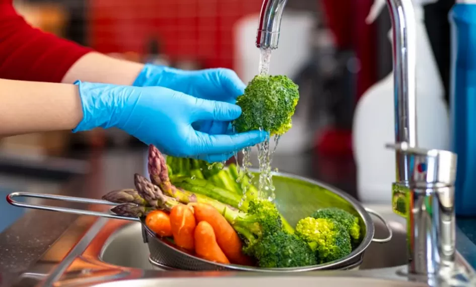 Manipulación de alimentos dos manos con guantes higienizando verduras bajo un grifo de cocina