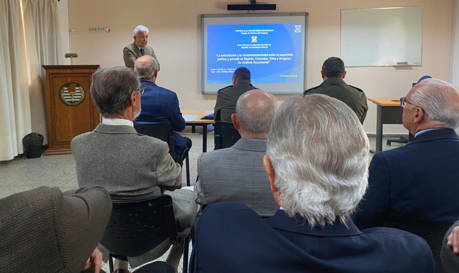Presentación de tesis: expositor de pie junto a pantalla, público lo escucha sentado. Presentación de tesis: expositor de pie junto a pantalla, público lo escucha sentado.