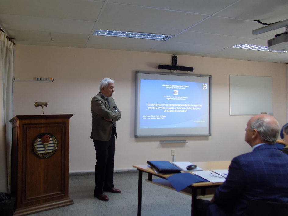 Presentación de tesis: expositor de pie junto a pantalla, público lo escucha sentado. Presentación de tesis: expositor de pie junto a pantalla, público lo escucha sentado.