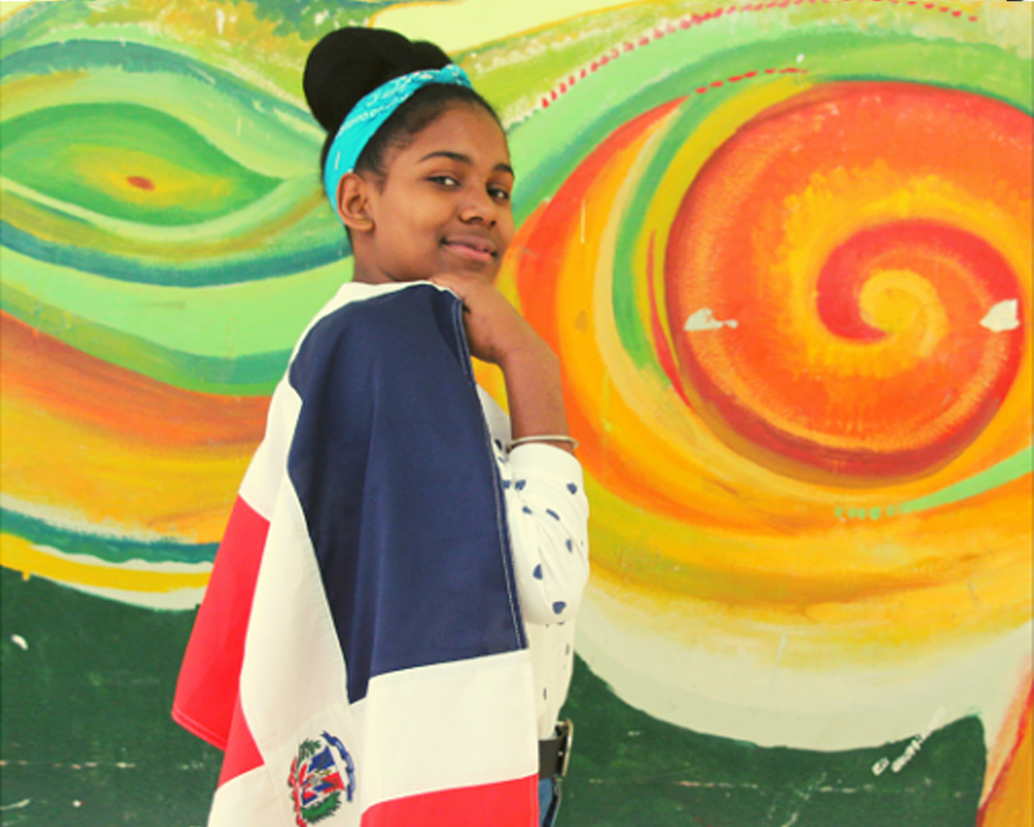 Muchacha joven envuelta en bandera de República Dominicana, fondo mural de colores.