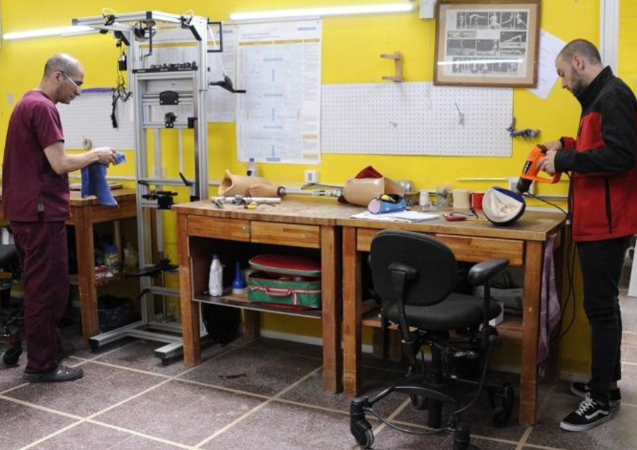 Dos personas trabajando en un taller de ortopedia. Sobre mesas de madera se ven herramientas y parte