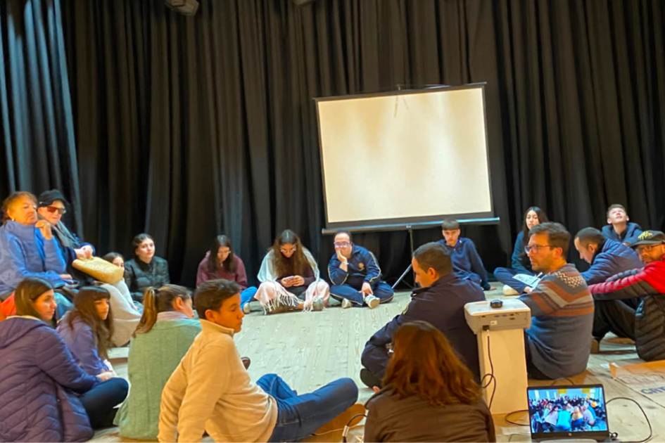 Personas participantes durante el encuentro Grupo de personas sentadas en círculo sobre un escenario, durante la actividad
