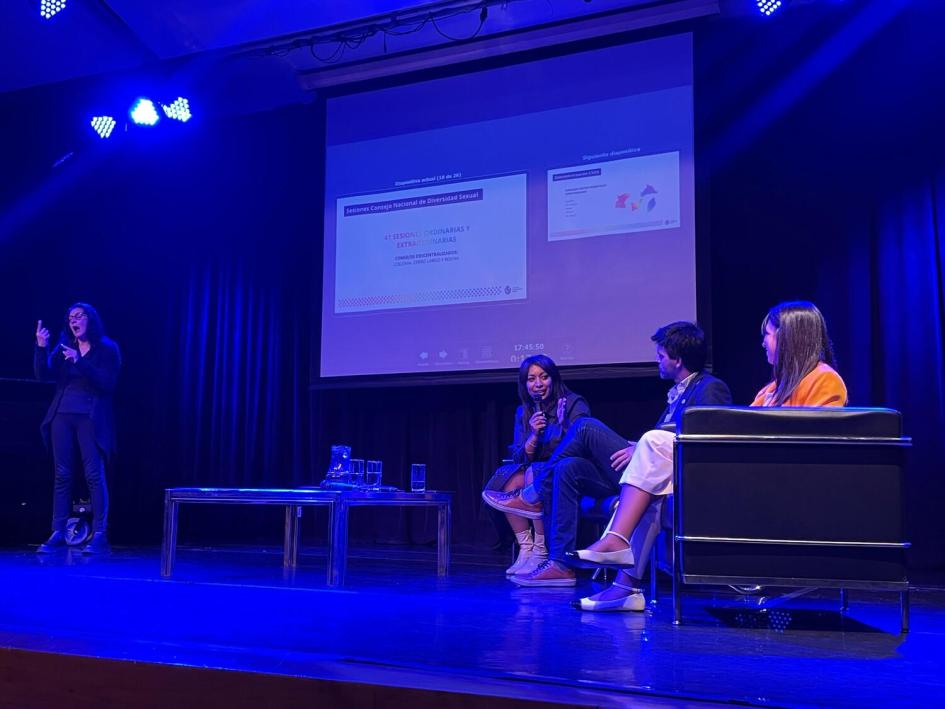 Autoridades ministeriales durante la presentación del Mes de la Diversidad Escenario, 3 autoridades sentadas en sillones, pantalla de fondo, intérprete de LSU de pie.
