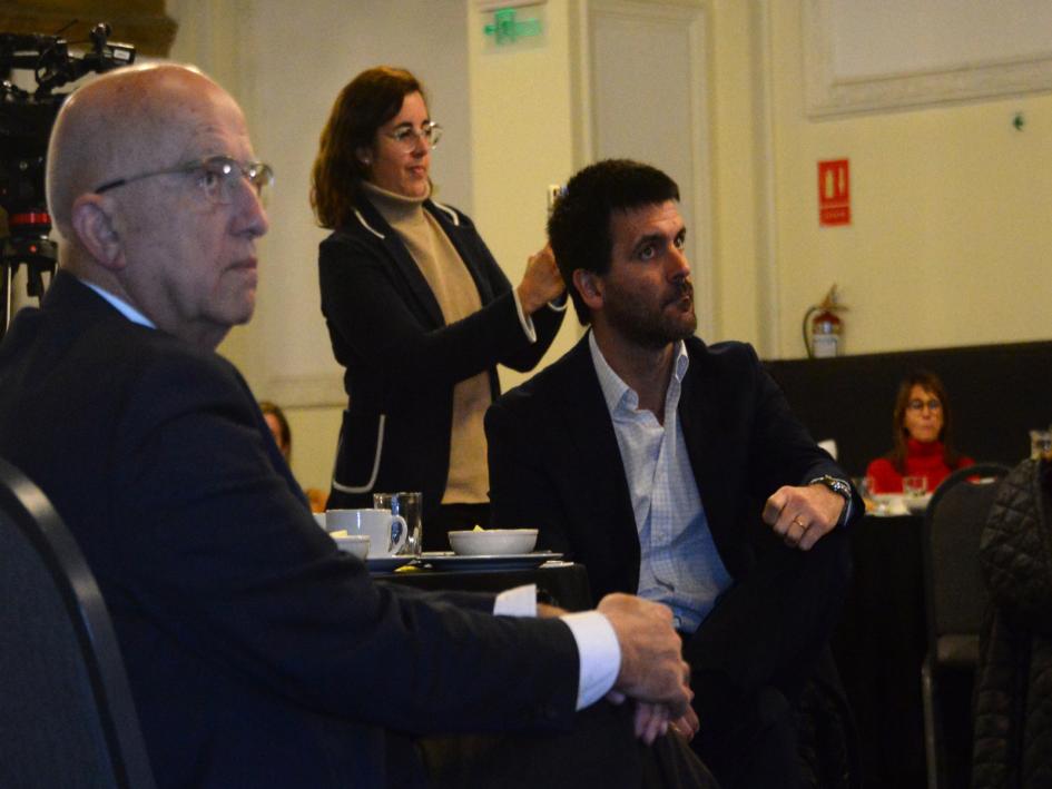 Ministro Alejandro Sciarra presente en el conversatorio. Foto: Presidencia de la República Sciarra sentado a la mesa junto a hombre, miran escenario. Detrás mujer saca fotos con celular.