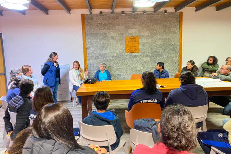 Personas participantes durante el encuentro Taller grupal en una sala con mesa larga, donde una mujer de pie habla al público.