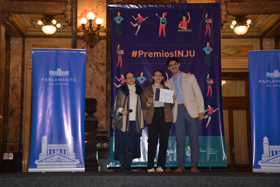 Entrega del premio a Antonella Pascual en la categoría Bienestar Animal Tres personas frente a los banners de Premios INJU en entrega de premio a categoría Bienestar Animal