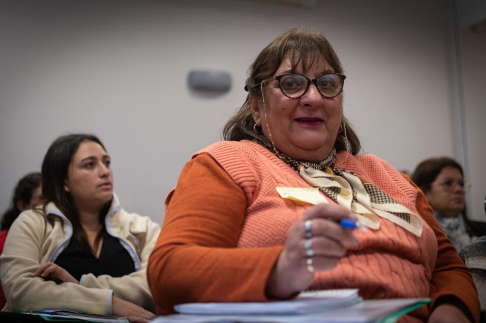 Mujer en primera plana que toma apuntes sonriendo en el fondo se ven mujeres participantes del curso