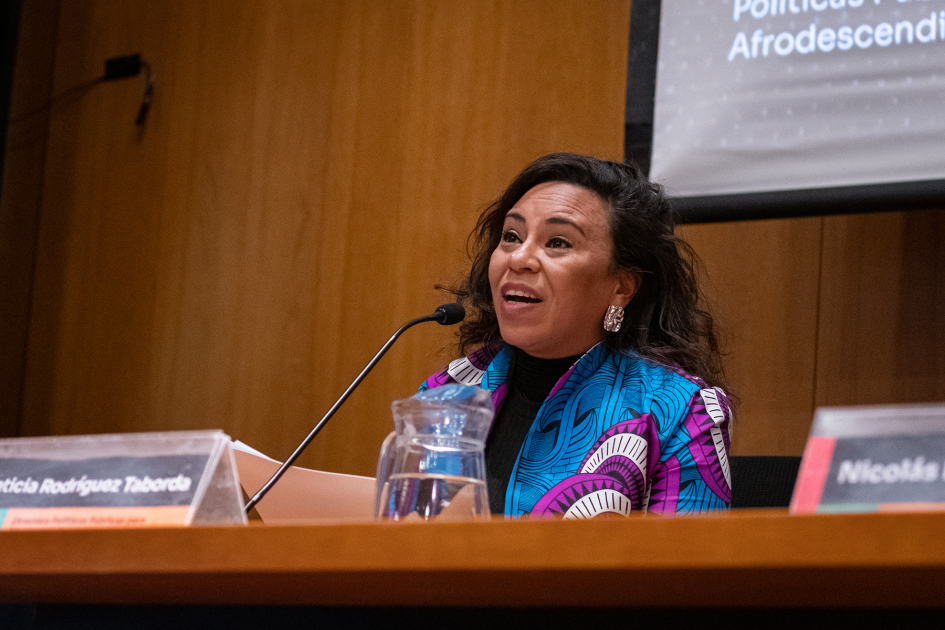 Directora de de la División de Promoción de Políticas Públicas para Afrodescendientes Mujer afro de mediana edad en la mesa de autoridades le habla al auditorio.