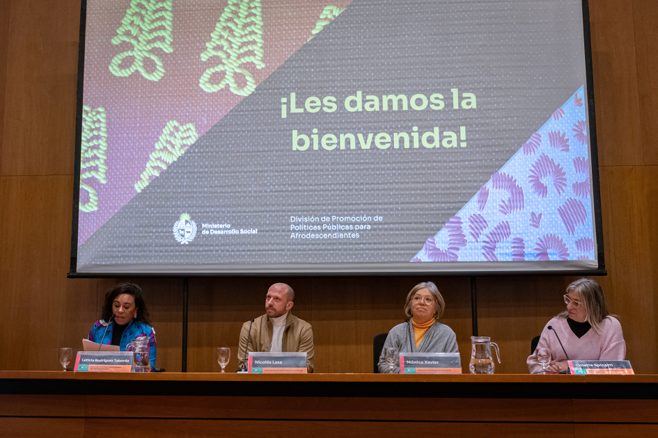 Mesa de autoridades en la bienvenida al lanzamiento del Mes de la Afrodescendencia Tres mujeres y un hombre en mesa de autoridades en escenario. Arriba pantalla de bienvenida.