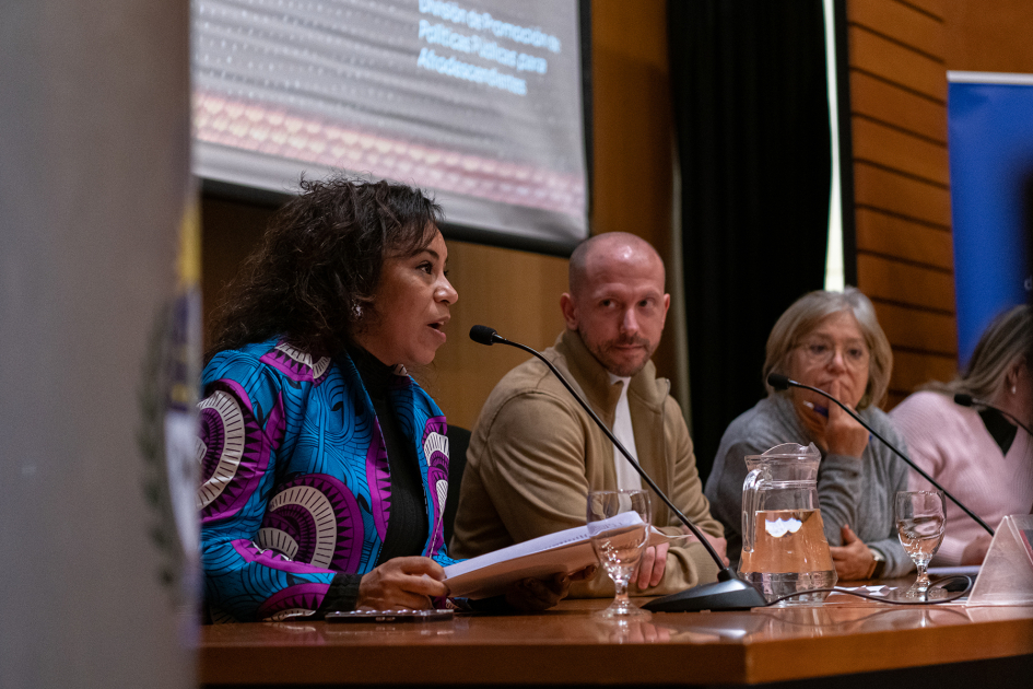 Leticia Rodríguez Taborda en su discurso de bienvenida y lanzamiento del mes. Rodríguez Taborda habla con carpeta en mano; Nicolás Lasa y Mónica Xavier en mesa de autoridades.