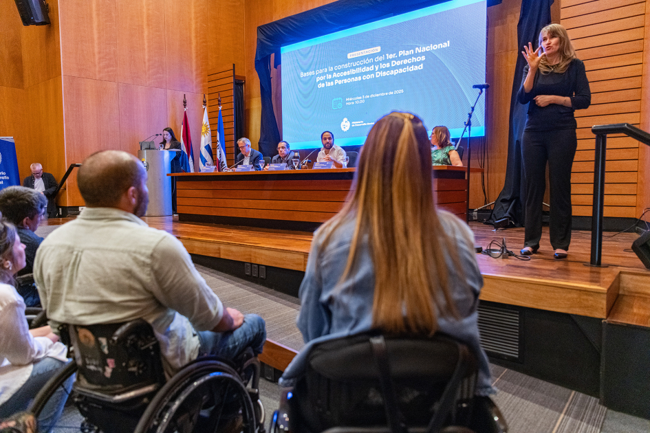 Plano frontal de la mesa, con toma de espaldas de personas en sillas de ruedas y foco en interprete 
