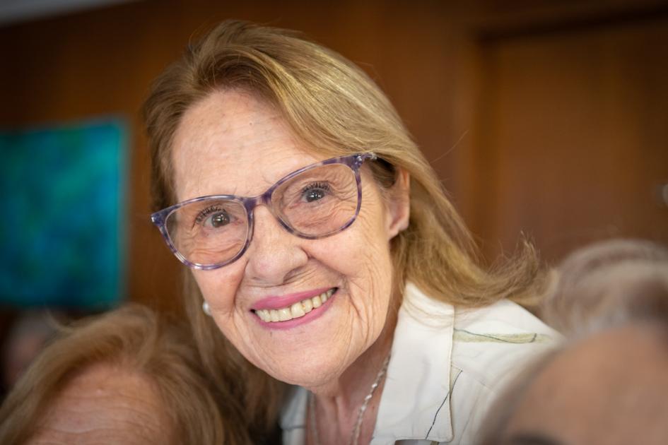 Público presente en el evento. Señora mayor, cabello largo claro, con lentes, mira y sonríe a la cámara.