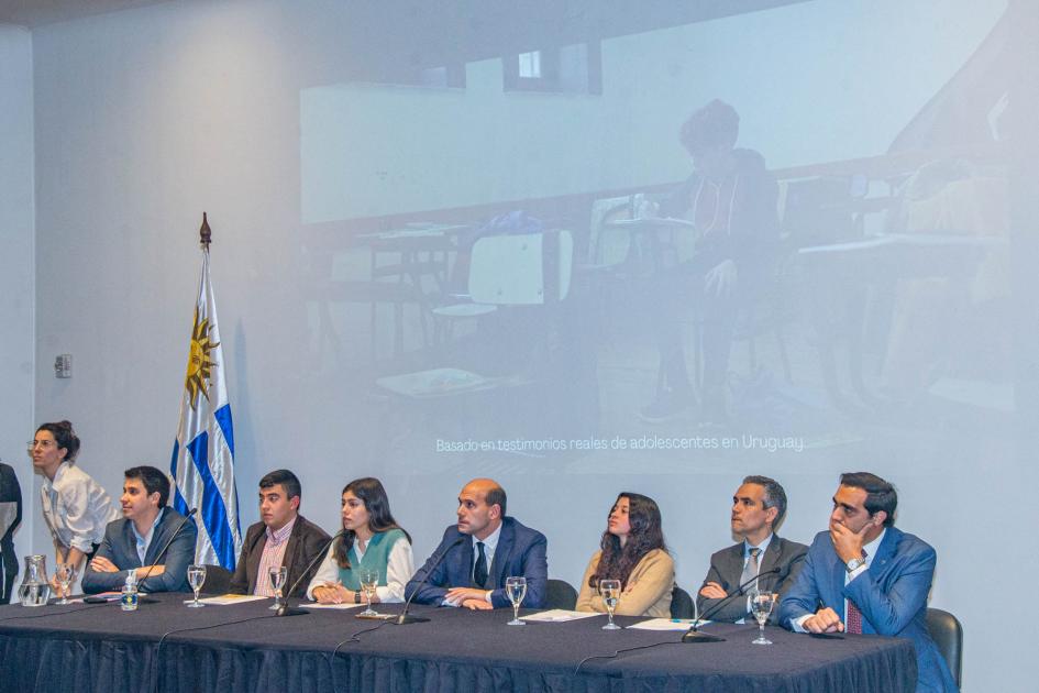 Mesa de oradores en lanzamiento de campaña #NiSilencioNiTabú” Mesa de oradores con el Pabellón Nacional de fondo y proyección de imagen de un adolescente