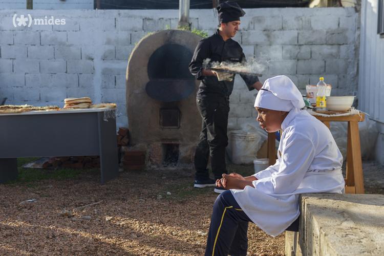 Maestro de pala saca pizza del horno de barro mientras estudiante está sentado en un murito