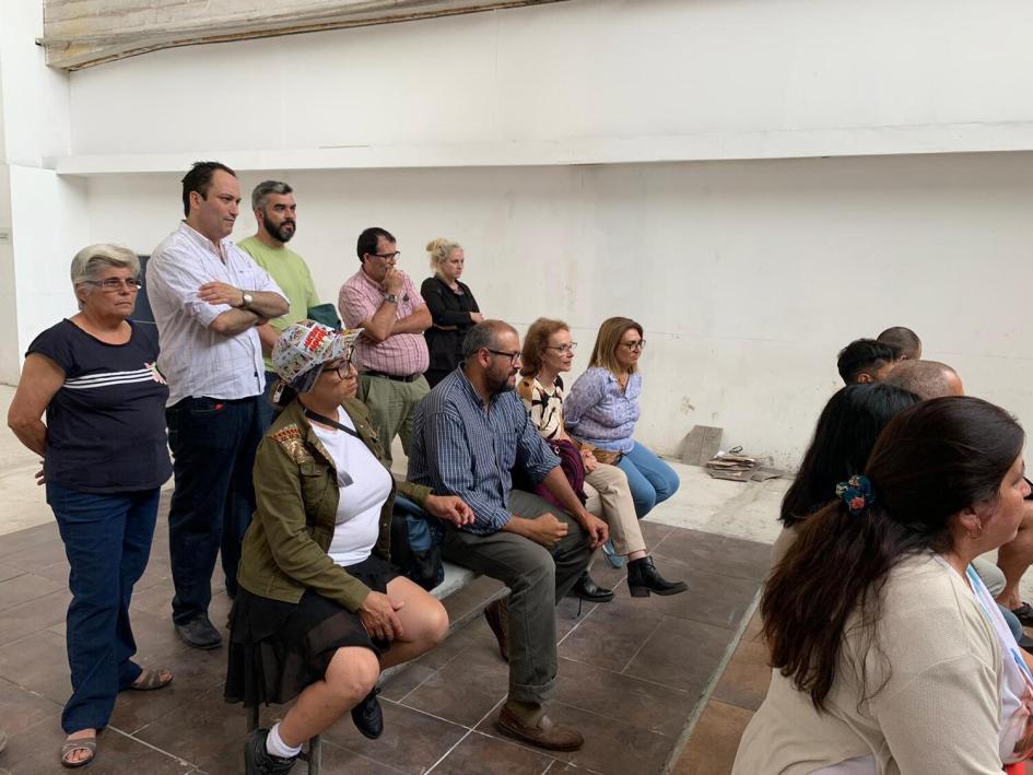 Docentes y alumnos esperan a ser llamados para recibir certificados. Alumnos sentados al fondo del salón, tras ellos de pie están los profesores.
