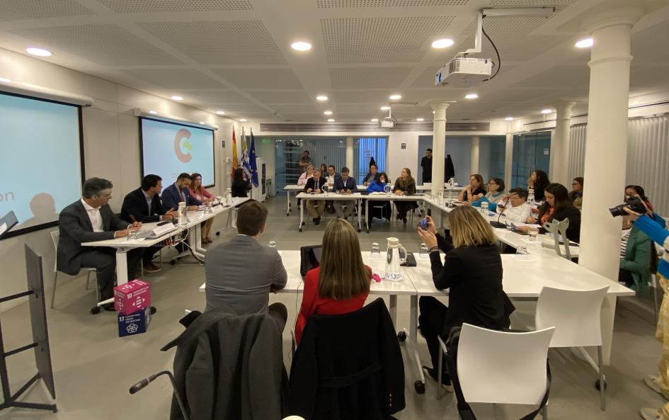 Sala de reuniones con varias mesas con disposición en "U". Se ven aproximadamente unas 20 personas.