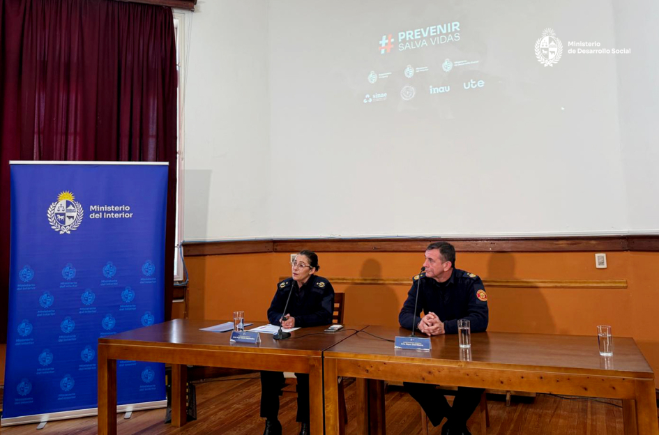 Valeria Vasconcellos y José Ribeiro en presentación de la campaña "Prevenir salva vidas". Mujer y hombre uniforme de bomberos, sentados a la mesa. Proyección en pared. En izquierda banner MI