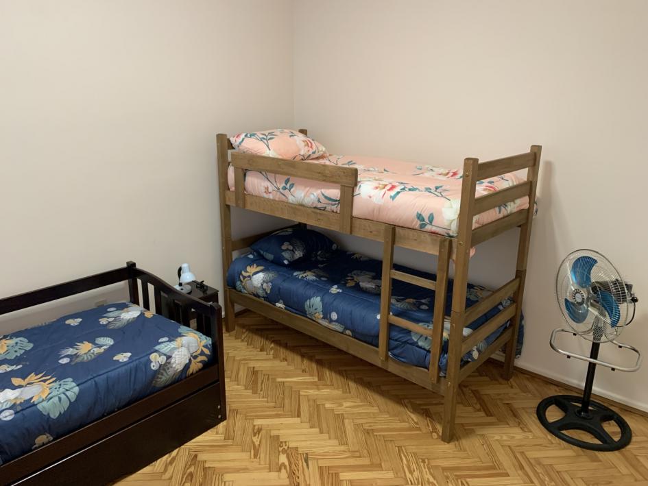 Otro de los dormitorios, cuchetas y cama marinera con acolchados azules, ventilador de pie.