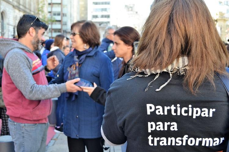 Participantes hablando con la ministra Arismendi de fondo y en primer plano, mujer con camiseta del programa de voluntariado que dice "Participar para transformar".