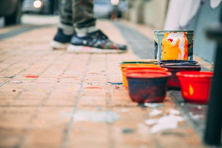 Tarros de pintura en la vereda y de fondo unos pies frente a un muro.