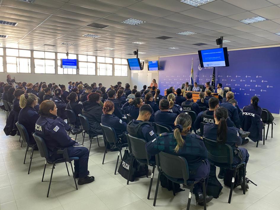 presentación cuaderno de denuncias accesible 3 auditorio conformado por 80 agentes policiales, vistos de atrás. en el fondo, la mesa de expositores