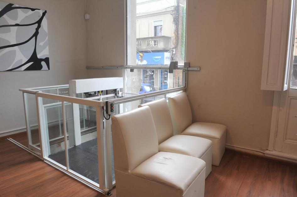 Plataforma vertical con elevador para silla de ruedas. Entrada a la casa, 3 sillones blancos, atrás una plataforma con elevador para las sillas de rueda.