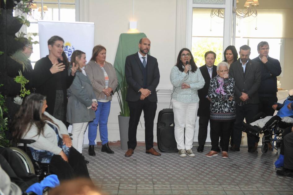 Personas de pie ubicadas en semicírculo, mujer habla en micrófono, habitación decorada con plantas.