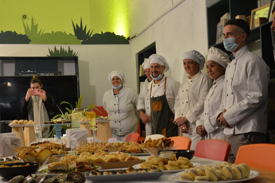 Parte del grupo de participantes que se capacitan en Gastronomía. Hombres y mujeres con uniforme entorno a una mesa repleta de productos panificados.