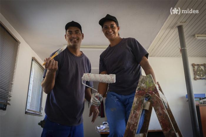 Participantes de la capacitación Dos persona miran y sonrien, la mujer que se encuentra del lado derecho tiene en su mano un rodillo de pintura y se sostiene en una escalera de dos hojas