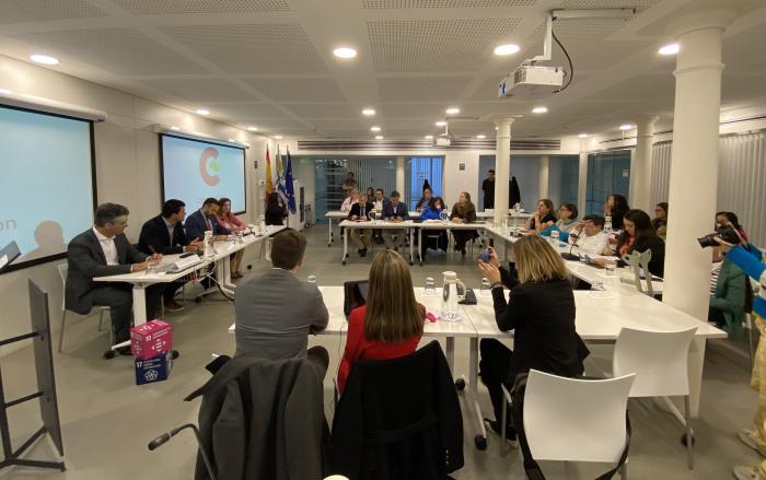 Sala de reuniones con varias mesas con disposición en "U". Se ven aproximadamente unas 20 personas.