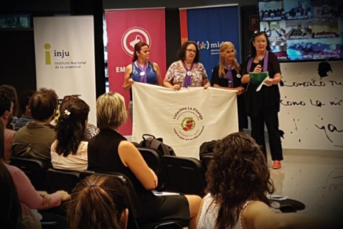 Presentación de los llamados a Fondos MIDES Cuatro mujere tienen en sus manos una bandera del colectivo Pitanga, frente a un auditorio de gente.