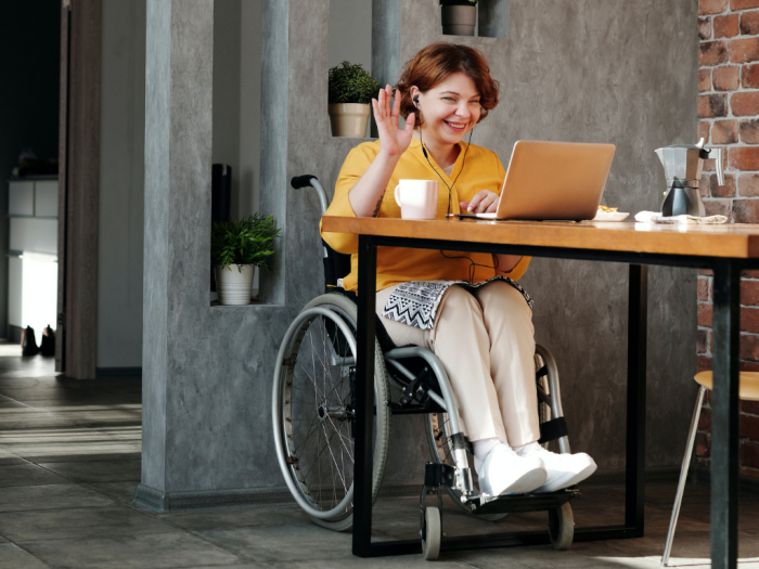 Persona en silla de ruedas sentada en una mesa frente a una computadora, saludando.