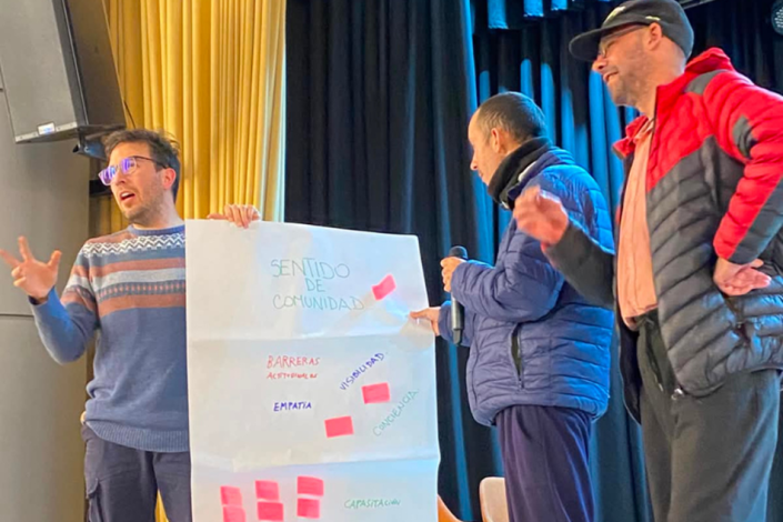 Personas participantes durante el encuentro Tres personas en un escenario; una sostiene un afiche que dice SENTIDO DE COMUNIDAD