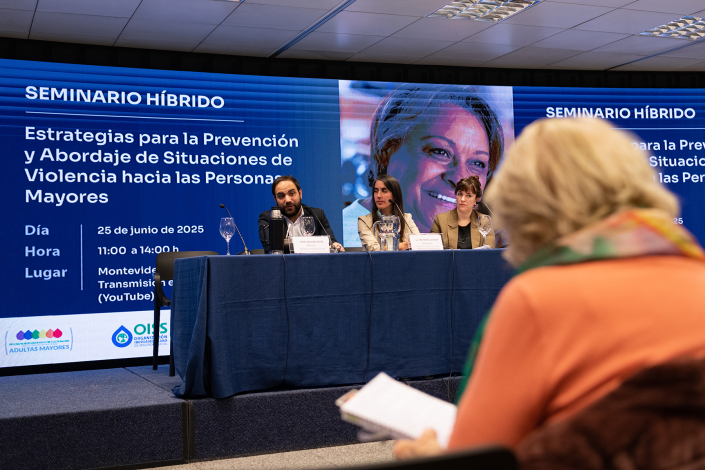Mujer mayor de espalda tomando apuntes, frente a ella autoridades del Mides en mesa de presentación.