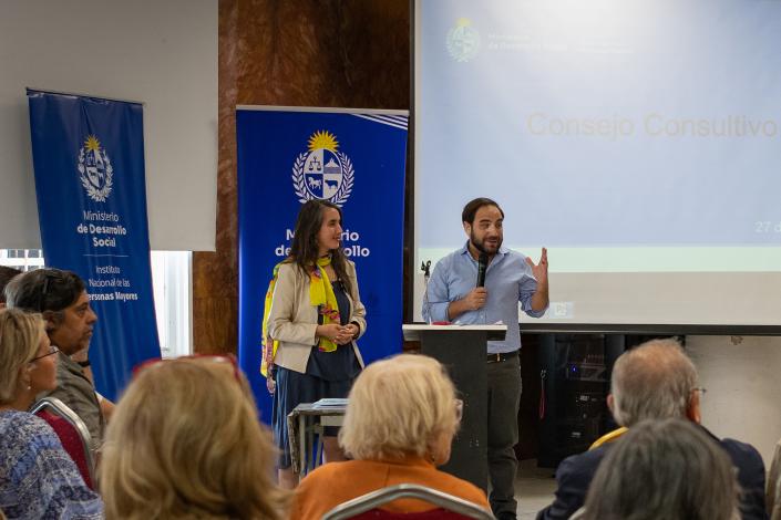 Personas mayores sentadas miran y escuchan a ministro Civila parado al frente junto a M. Larzábal.