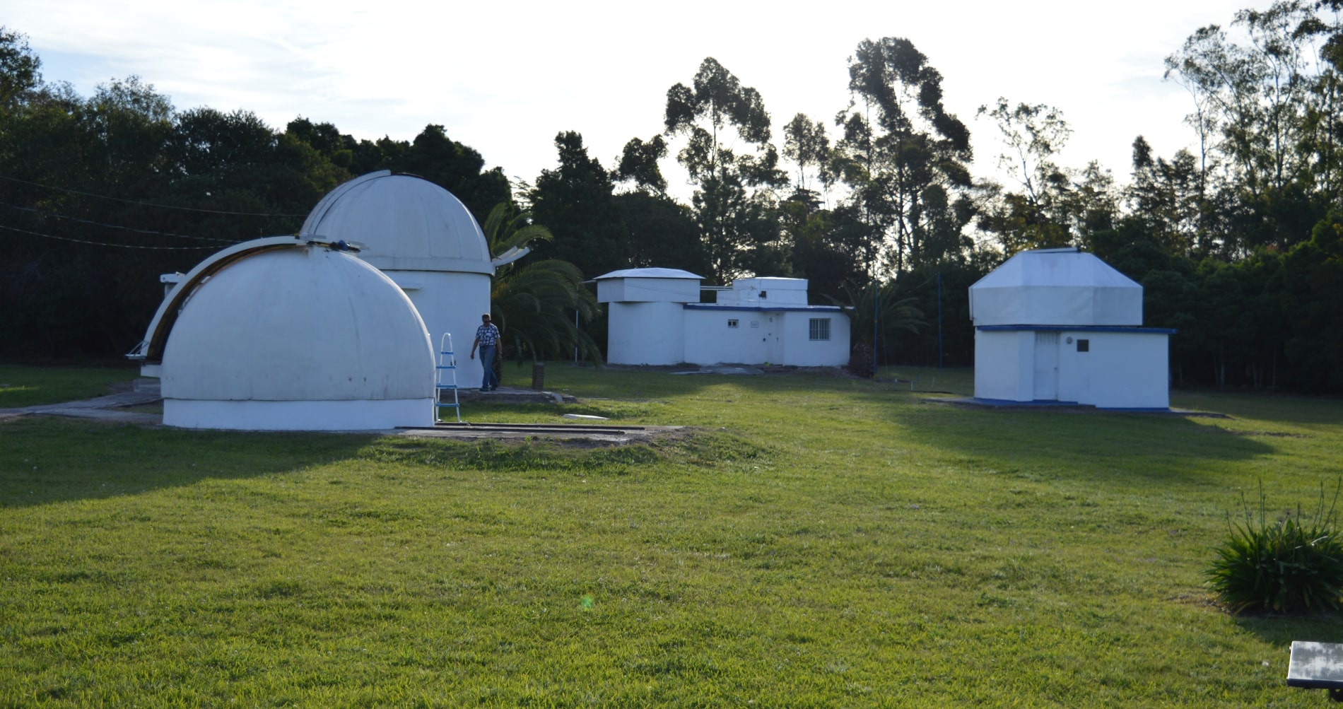 Observatorios astronómicos blancos en un predio de césped verde rodeado de árboles bajo un cielo claro.