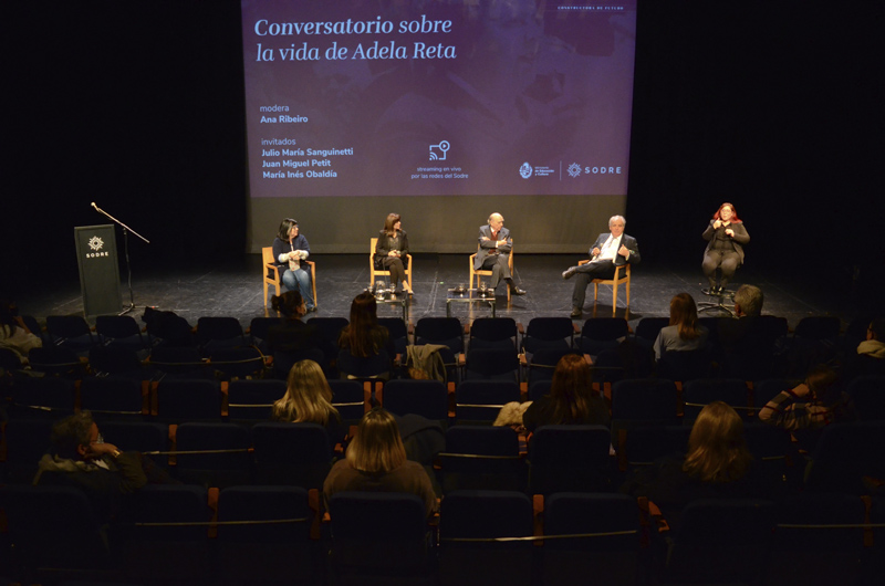 Público en sala Hugo Balzo y en el fondo se puede ver el escenario con las personas que participaron del conversatorio