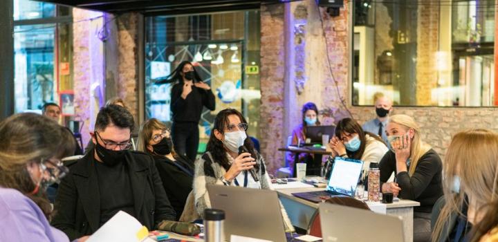 Participantes sentados y una de ellas está con un micrófono hablando