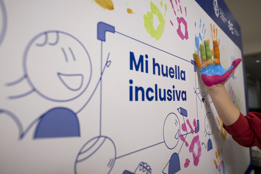Mano pintada de vivos colores (verde, naranja, azul, rosa) cerca del lema "Mi huella inclusiva" en una pared con huellas